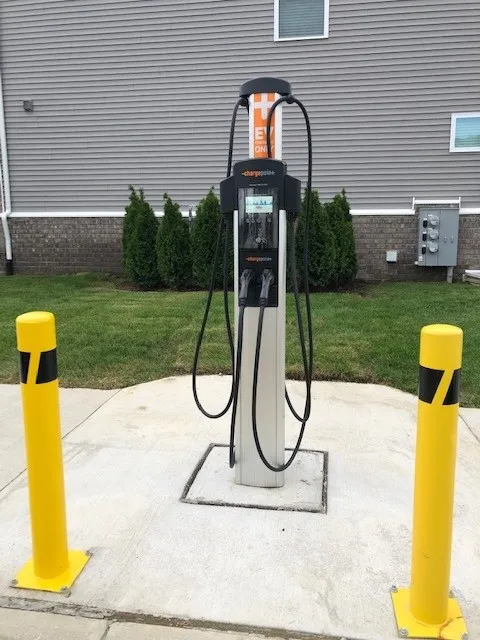 Modern electric vehicle charging station with sleek design, surrounded by well-maintained landscaping and protective yellow bollards.