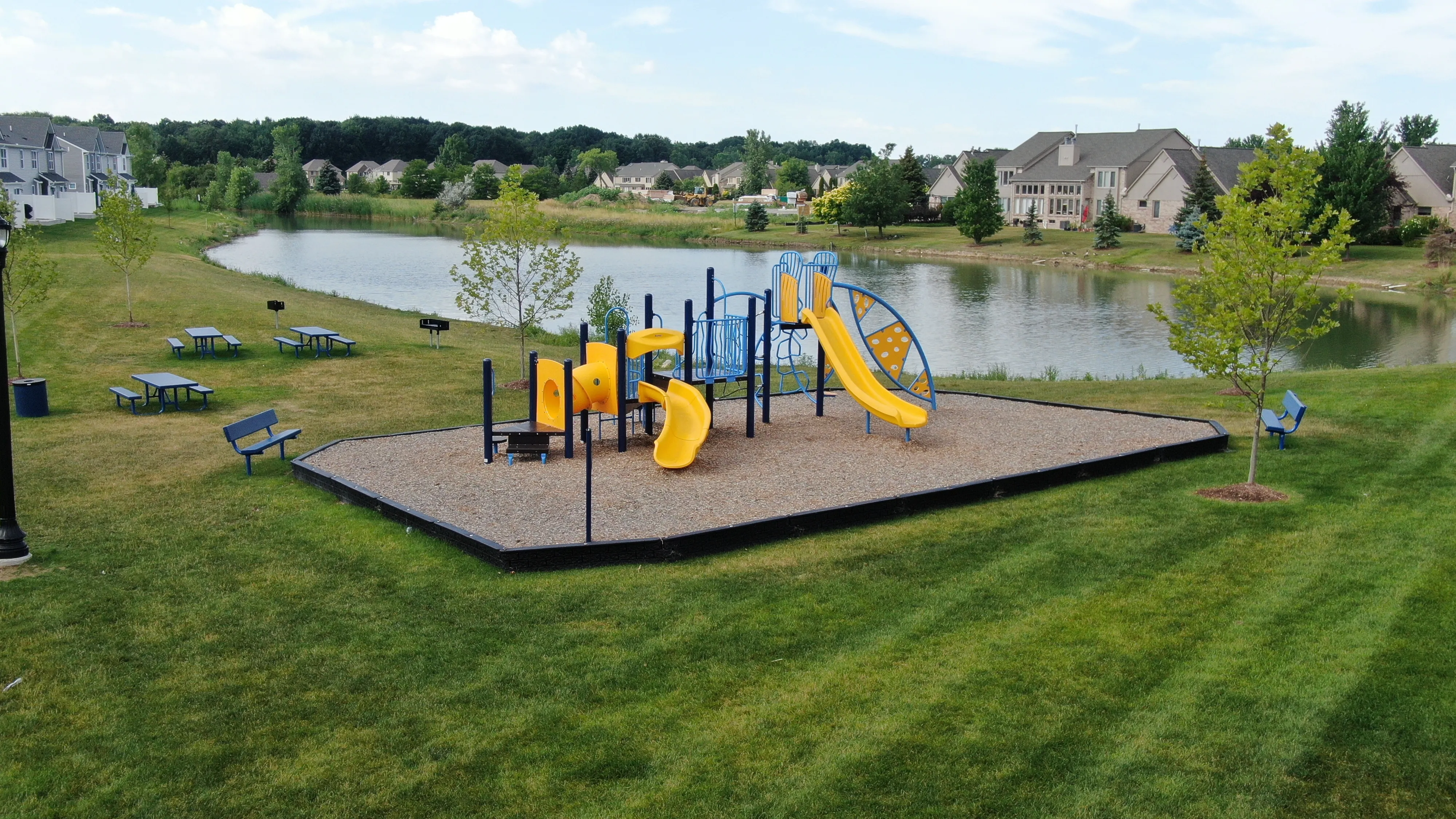 Charming playground with colorful slides and climbing structures in a tranquil lakeside community park, surrounded by lush greenery and picnic tables.