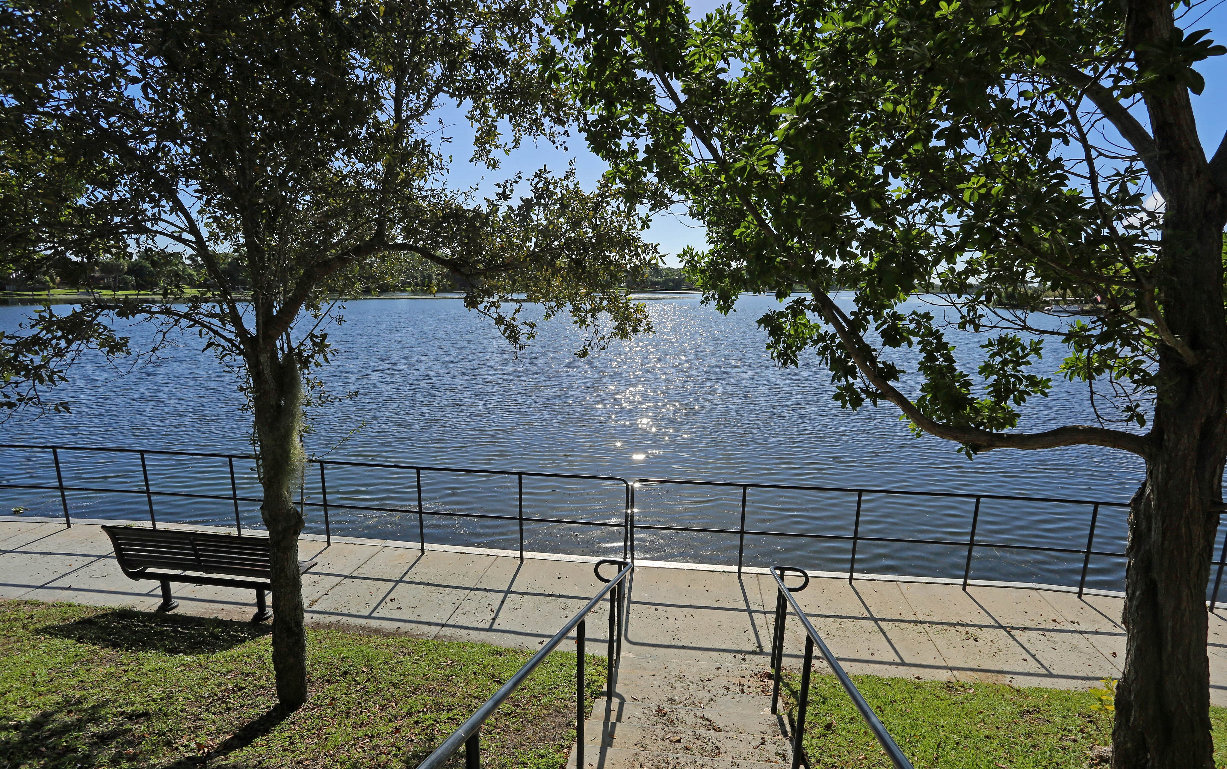 Lakeside walkway with a bench, iron railing, and stairway under tree shade overlooking calm water.