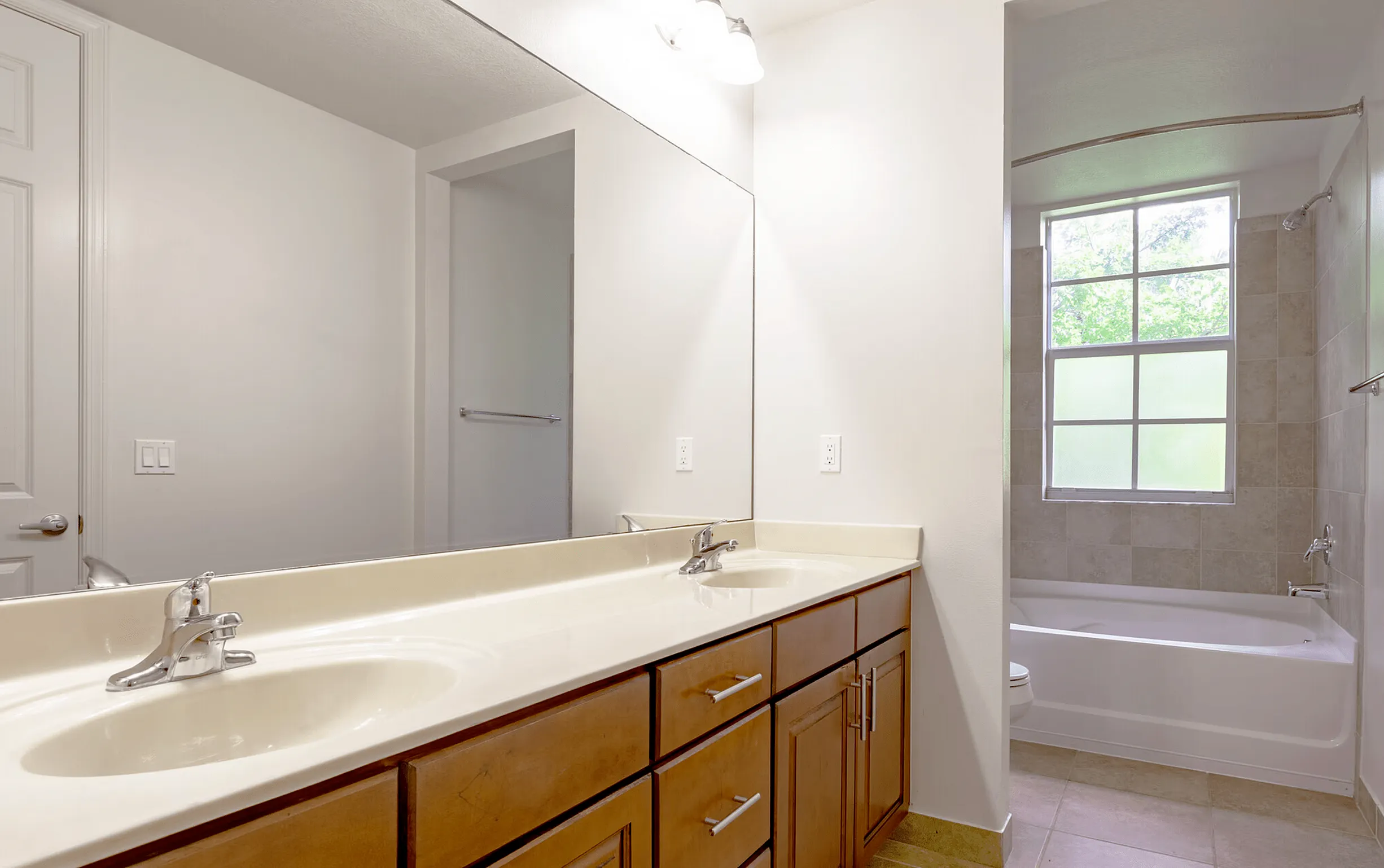 Bathroom with dual-sink vanity, large wall mirror, and bathtub with tiled surround beneath a frosted window.