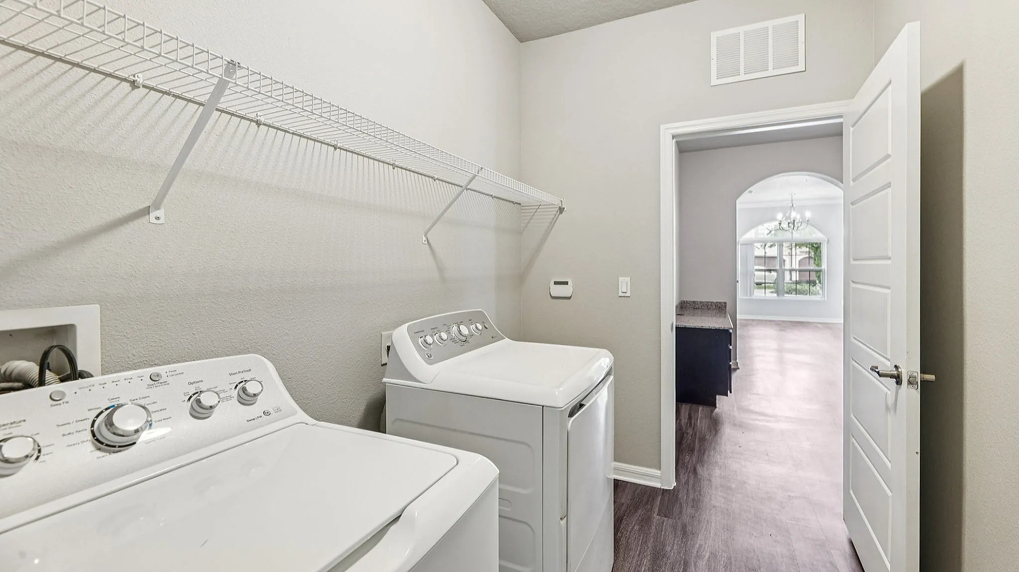 Modern laundry room with sleek white washer and dryer, featuring open shelving and view into sunlit living area with large window.