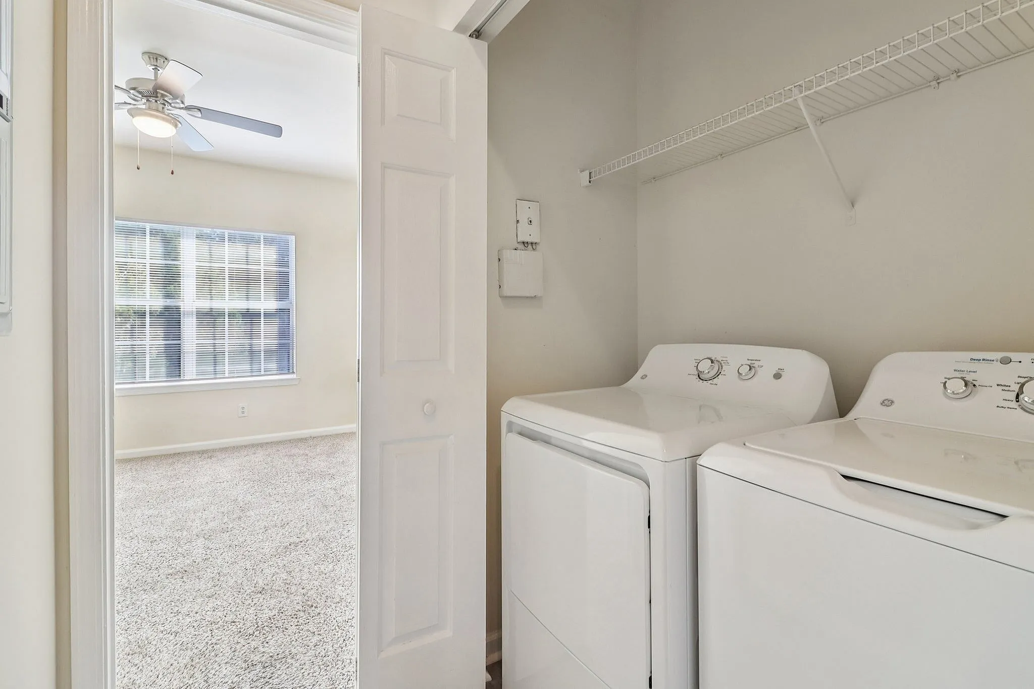 Bright laundry room with modern washer and dryer, adjacent to a sunlit room with carpet flooring and large window.