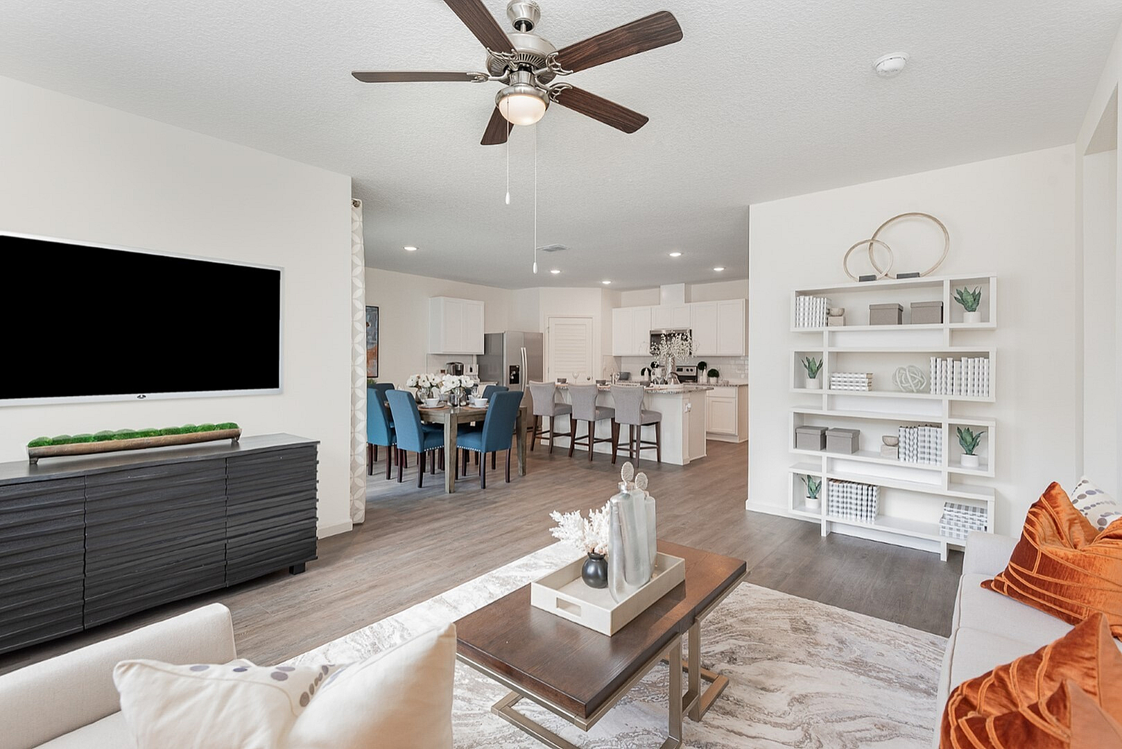 Inviting open-concept living area featuring a modern kitchen, elegant dining space with blue chairs, and a cozy living room with stylish decor and a ceiling fan.