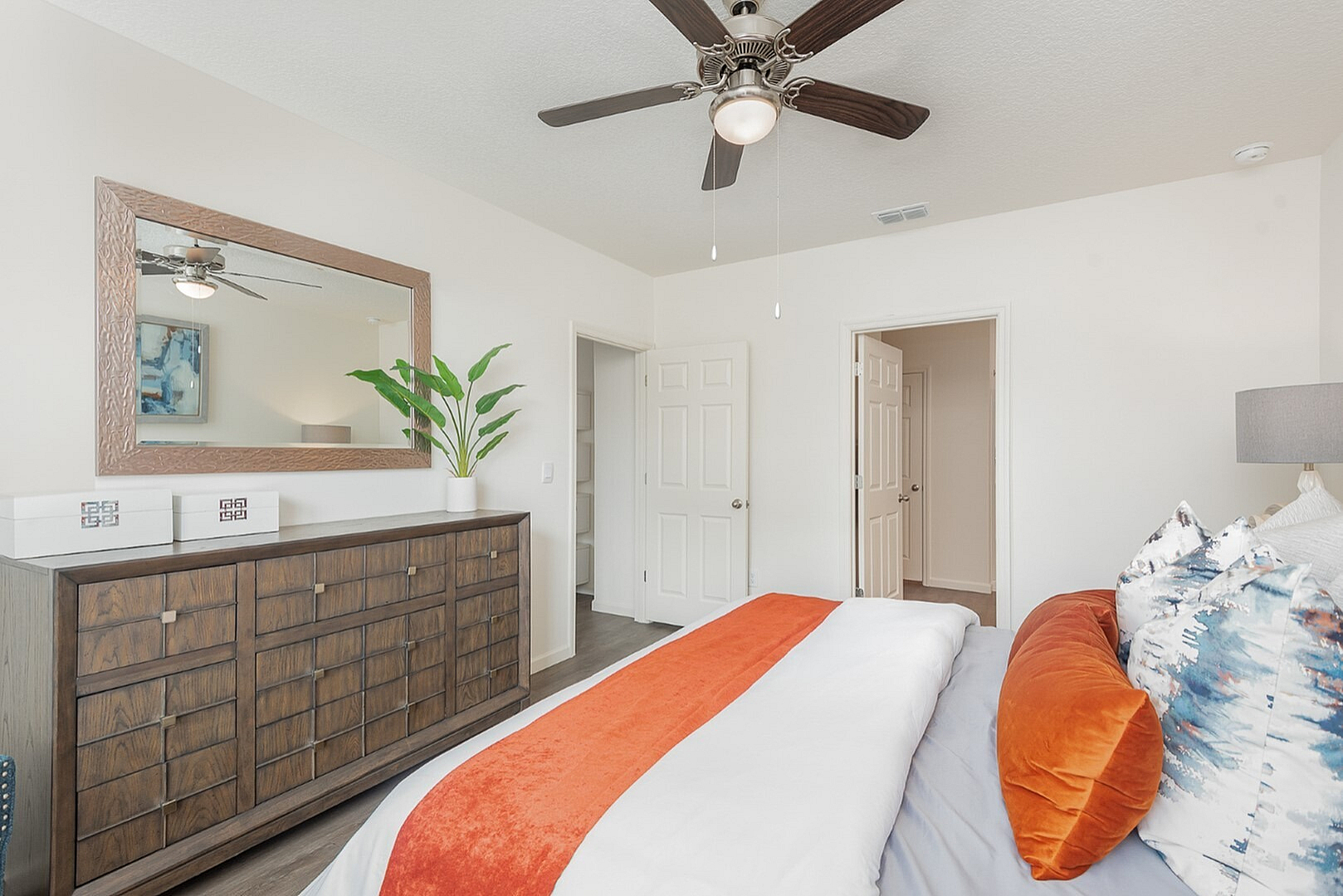 Chic bedroom with modern decor, featuring a stylish wooden dresser, vibrant pillows, and a ceiling fan, offering a cozy and inviting atmosphere.