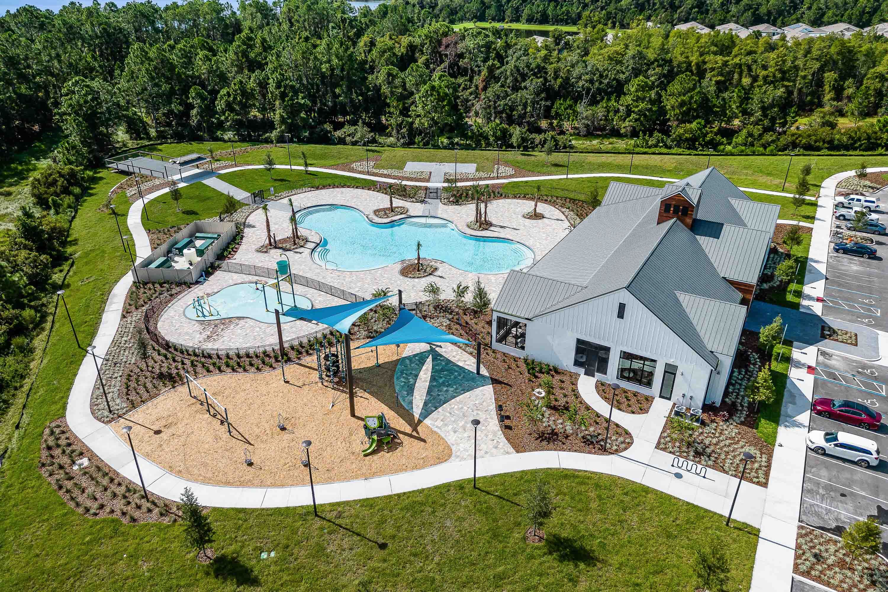 Aerial view of a luxurious community center with a uniquely shaped swimming pool, modern playground, and beautifully landscaped surroundings set against a lush green backdrop.