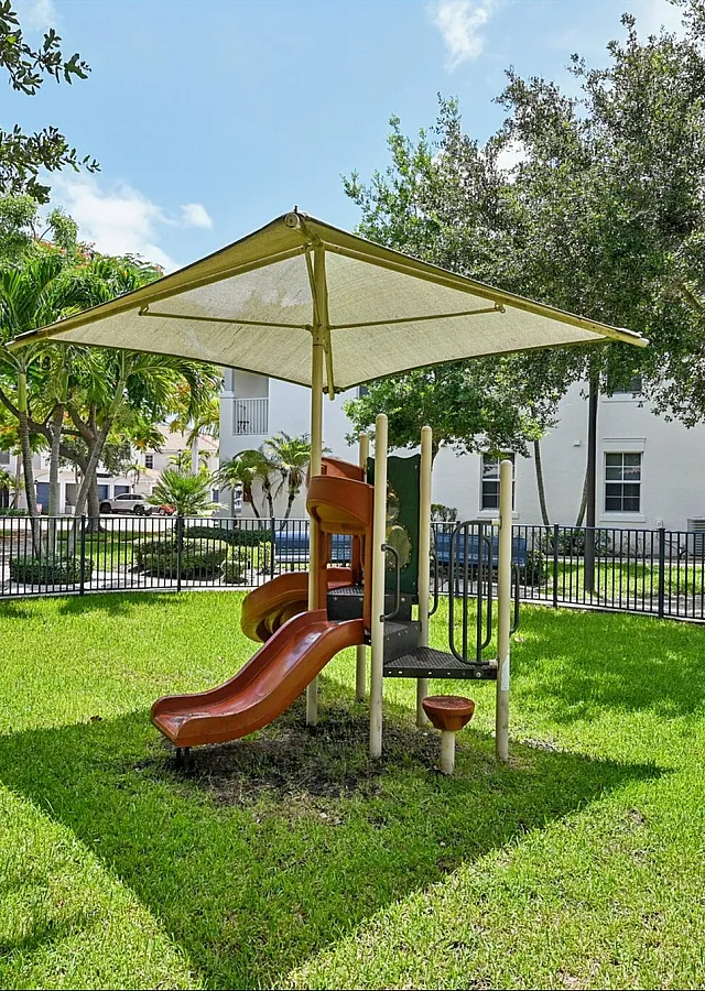 Charming apartment complex courtyard featuring a shaded playground with lush greenery and palm trees.