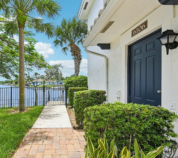 Charming lakeside entrance with lush greenery and a paved path leading to a serene water view under clear blue skies.
