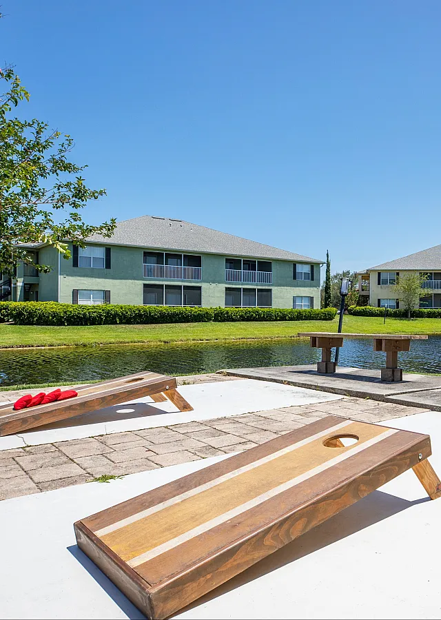 Sunny outdoor recreation area in a serene apartment setting with corn hole boards, picnic benches, and a tranquil water view.