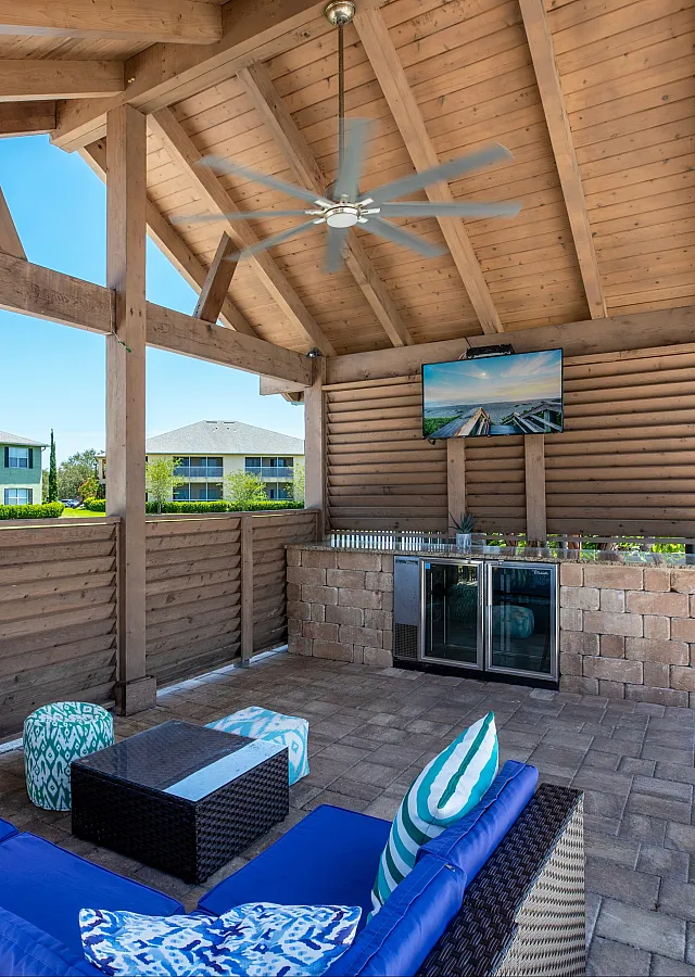 Charming outdoor patio with a cozy rattan sofa set, vibrant cushions, and a large TV under a rustic wooden pergola.