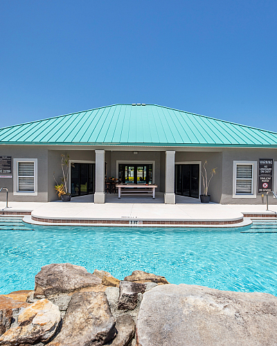 Inviting outdoor pool with crystal-clear water, surrounded by tropical plants and featuring a modern clubhouse with a vibrant teal roof.