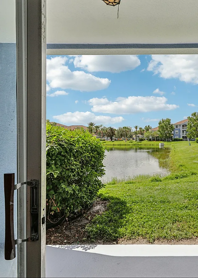 Inviting patio view showcasing lush greenery, a serene pond, and charming residential buildings under a bright blue sky.