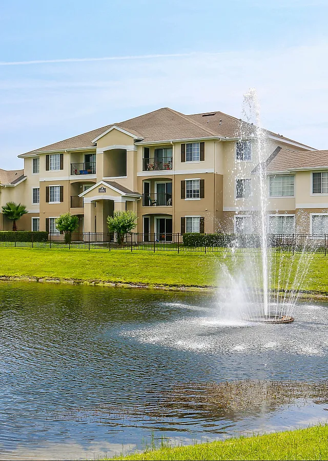 Elegant apartment complex with landscaped lawns and a serene pond featuring a fountain, offering peaceful lakeside living.