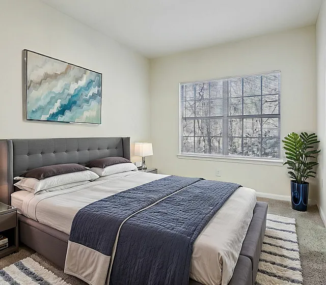Inviting bedroom with stylish grey upholstered bed, elegant artwork, and large window offering natural light.