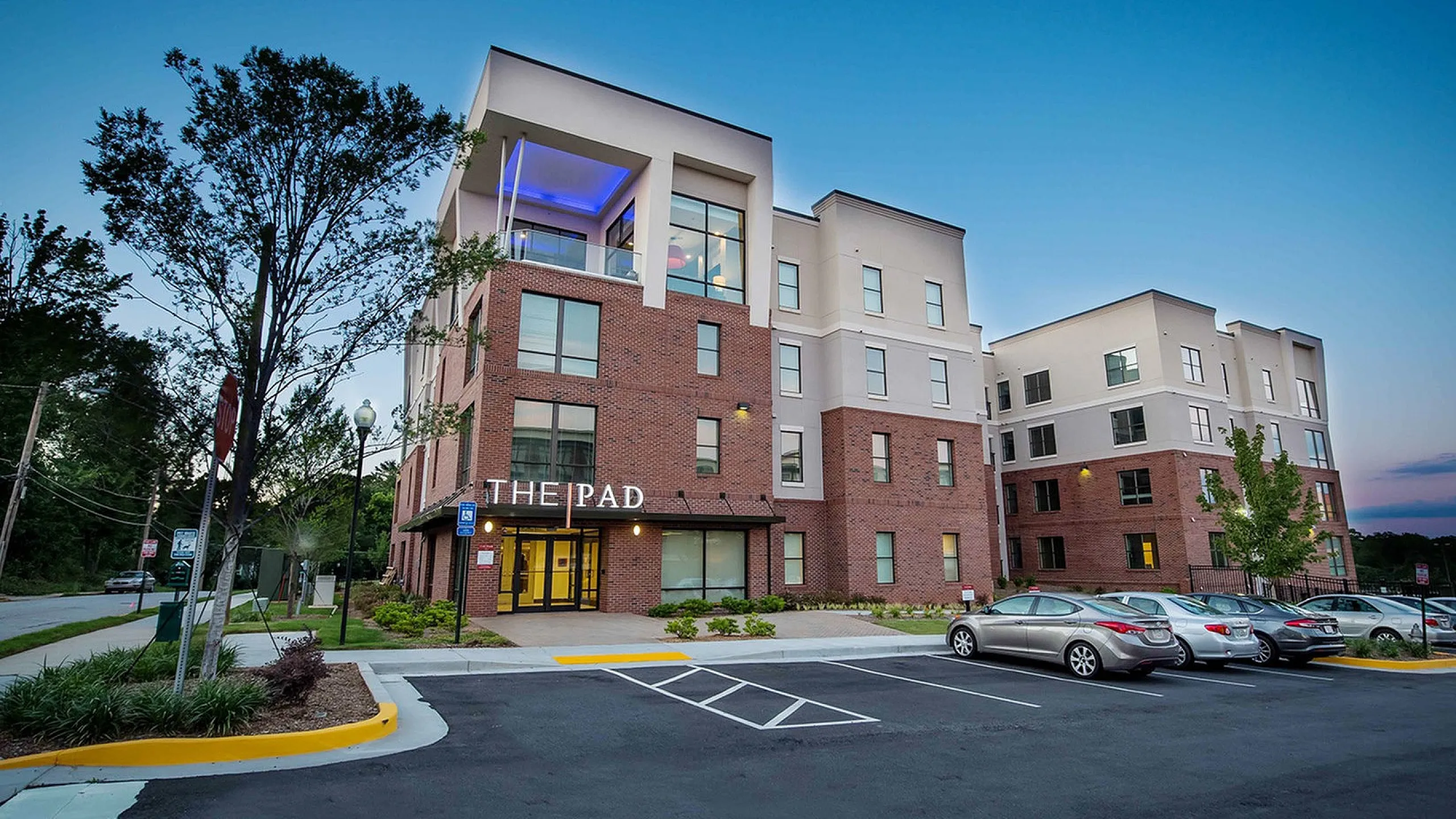 Modern apartment complex with stylish architecture and reserved parking, surrounded by lush greenery at dusk.