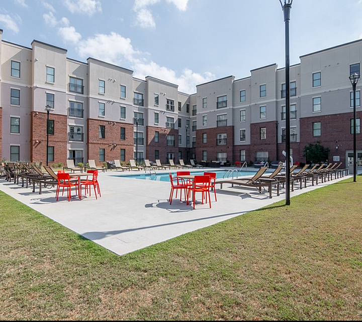 Modern apartment complex with a beautiful outdoor pool and lounge area, featuring vibrant red seating and sun loungers under a clear blue sky.