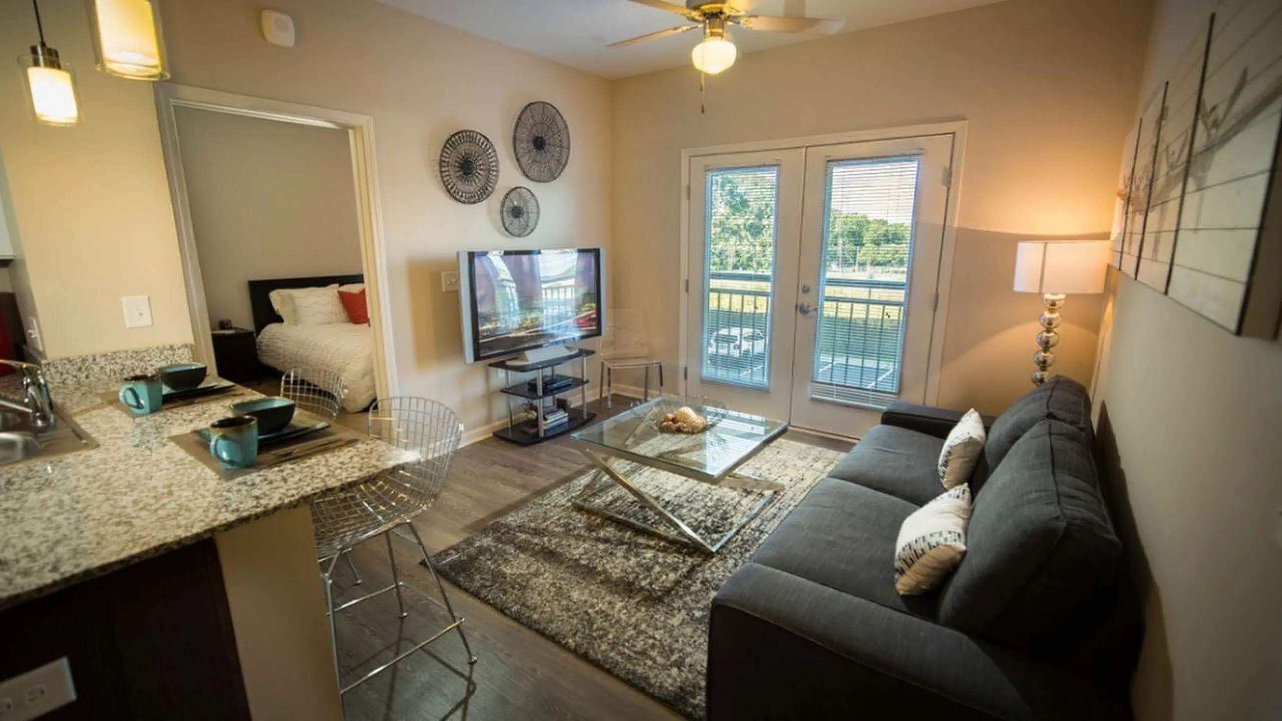 Chic living room with a cozy sofa, glass coffee table, and flat-screen TV, complemented by a granite kitchen counter and vibrant bedroom in the background.