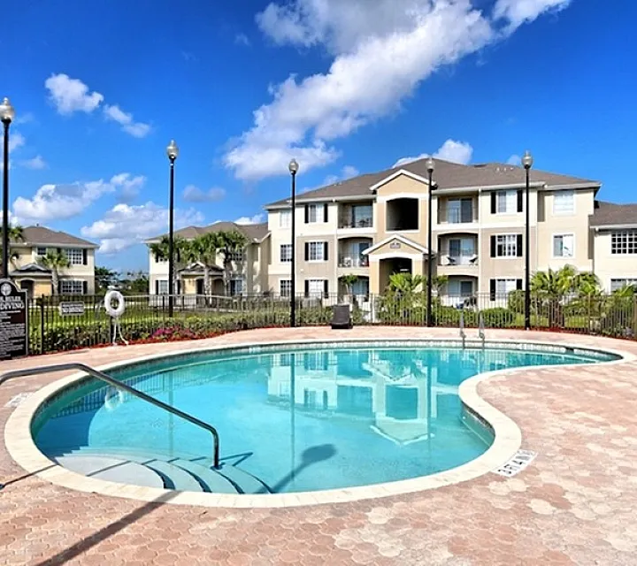 Inviting apartment complex with a crystal-clear swimming pool, beautiful landscaping, and elegant multi-story buildings under a bright blue sky.