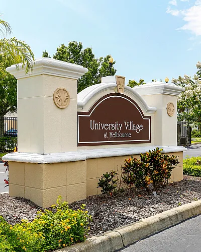 Inviting entrance of University Village at Melbourne with elegant signage surrounded by lush greenery and palm trees, promising serene and stylish living.