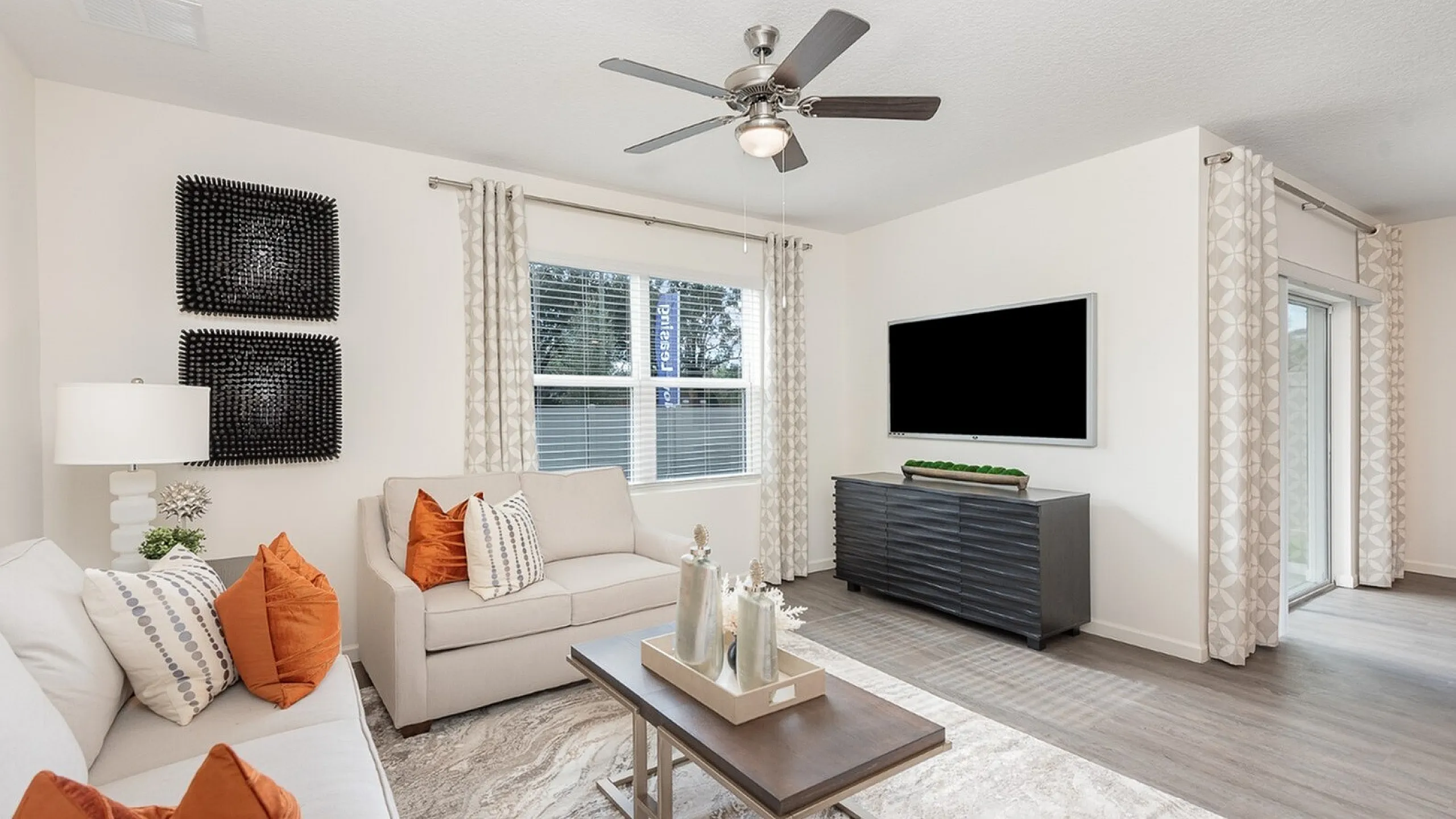 Bright and inviting living room with modern decor, featuring plush sofas adorned with vibrant orange pillows, a stylish coffee table, and a large flat-screen TV, perfect for relaxing or entertaining.