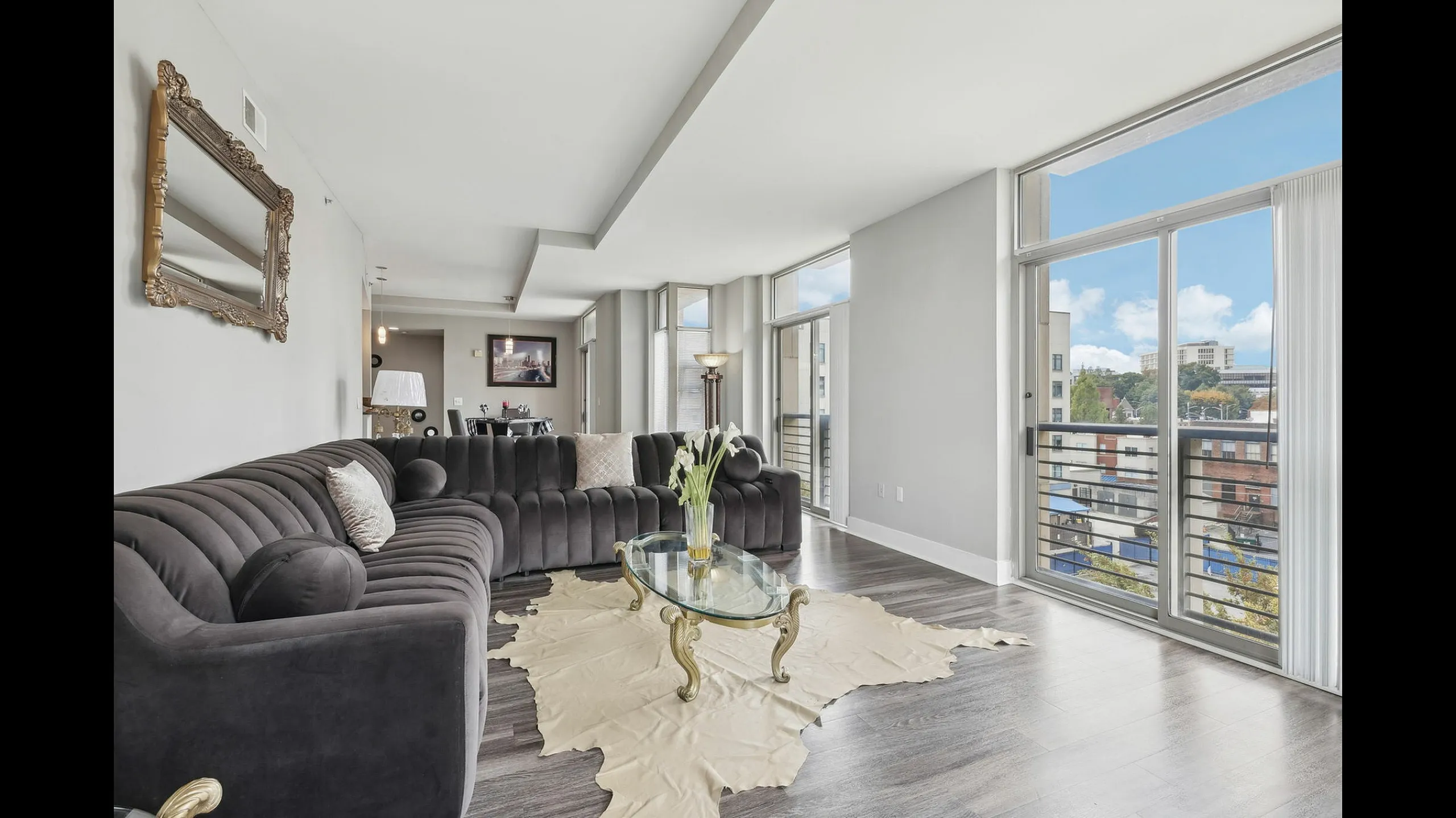 Elegant modern living room with large windows offering a cityscape view, featuring a plush gray sectional sofa and a stylish glass coffee table on a chic area rug.