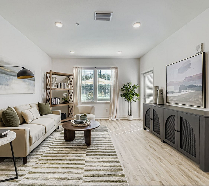 Elegant modern living room with a cozy beige sofa, stylish artwork, and a sleek dark wood cabinet. Sunlit through large window, featuring tasteful decor and a plush rug for a warm, inviting ambiance.