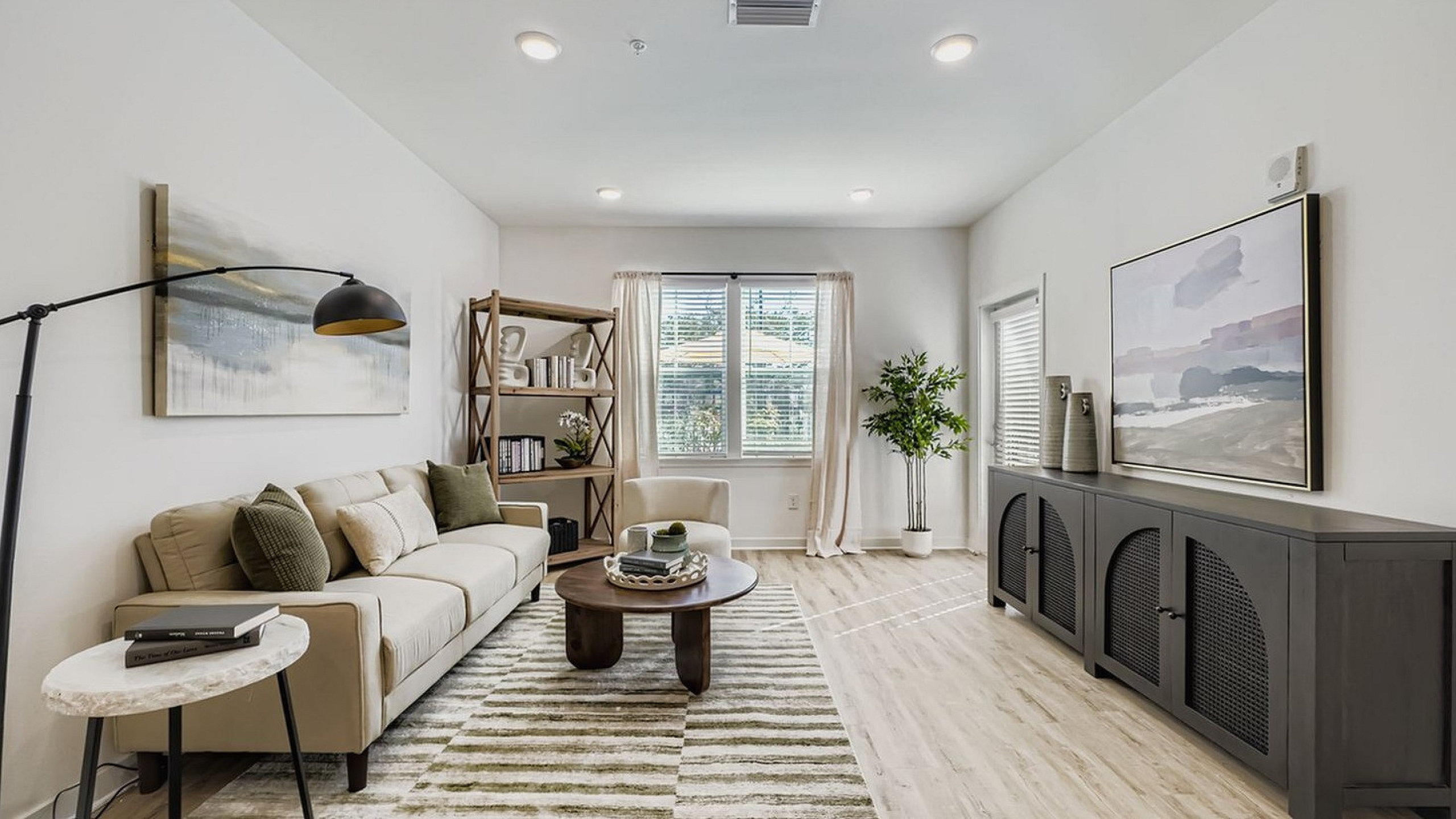 Bright and inviting living room with modern decor, featuring a cozy beige sofa, stylish floor lamp, and elegant bookshelf. Natural light streams through large windows, illuminating the airy space.