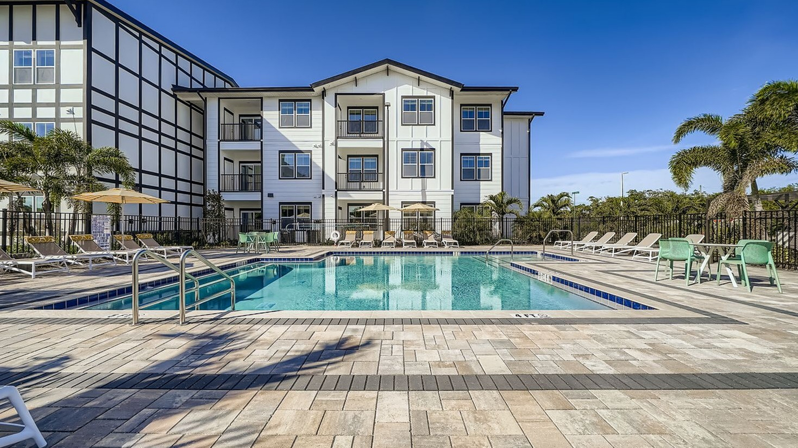 Contemporary apartment complex with a luxurious outdoor pool, surrounded by sun loungers and palm trees, under a clear blue sky.