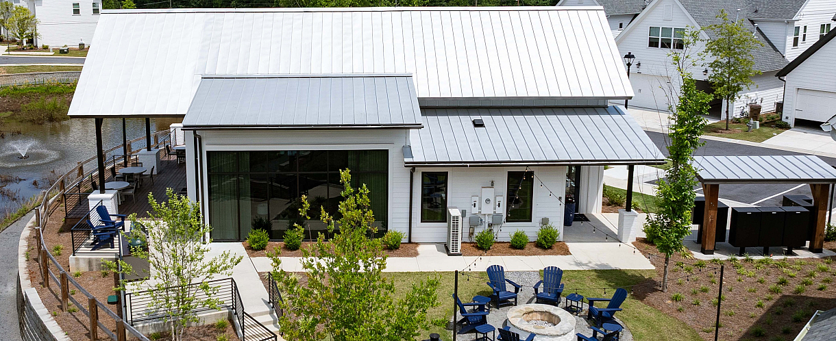 Charming community view featuring a modern clubhouse by a tranquil pond, surrounded by lush greenery and cozy seating areas with a fire pit.