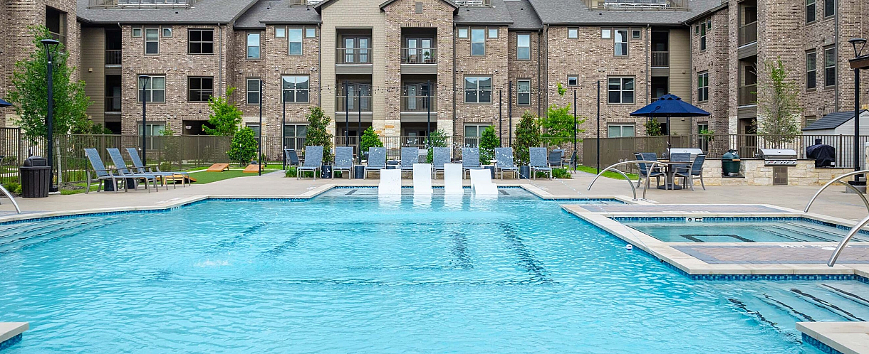 Inviting apartment complex with a luxurious outdoor swimming pool, sun loungers, and stylish tables under umbrellas amidst lush greenery.