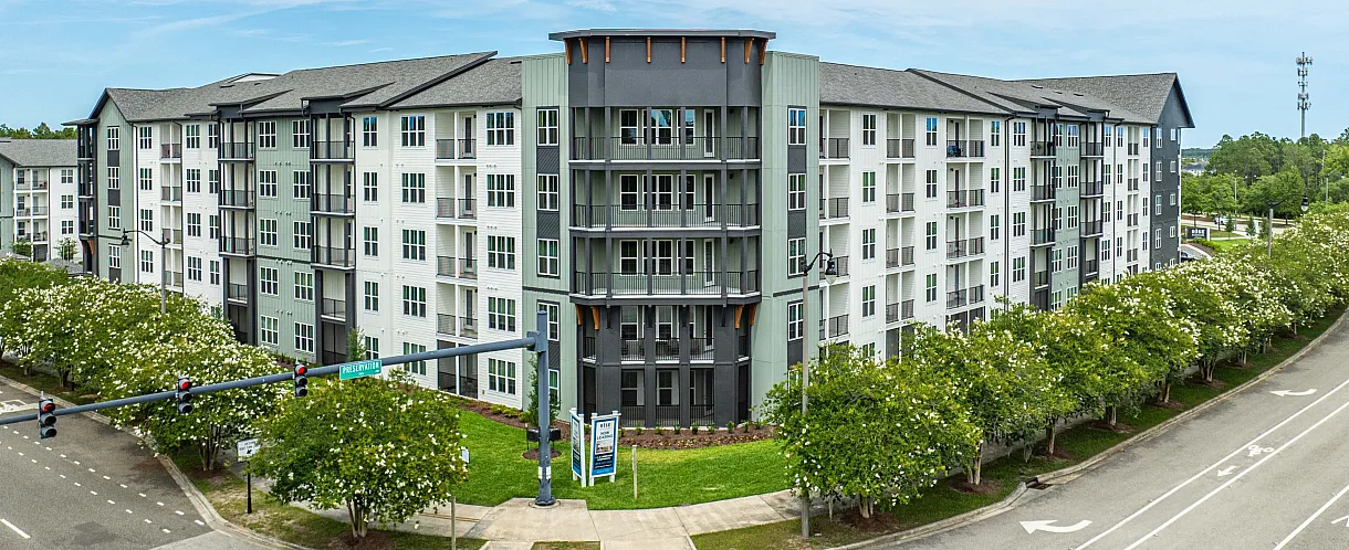 Modern multi-story apartment building with sleek architectural design, nestled at a street corner surrounded by lush greenery and bright skies.