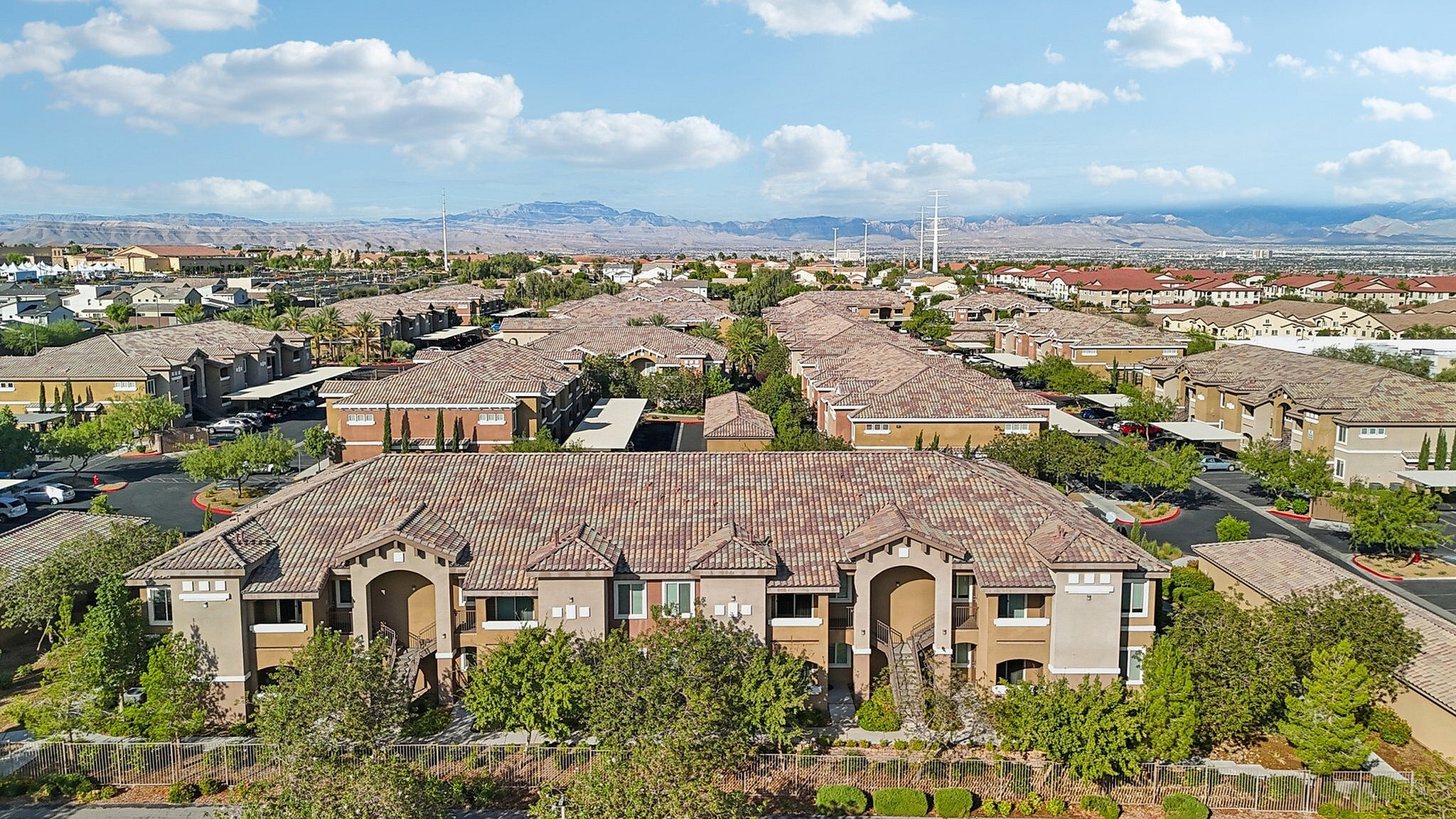 Aerial view of a charming suburban neighborhood with tile-roofed homes, tree-lined streets, and scenic mountain backdrop under a bright blue sky.