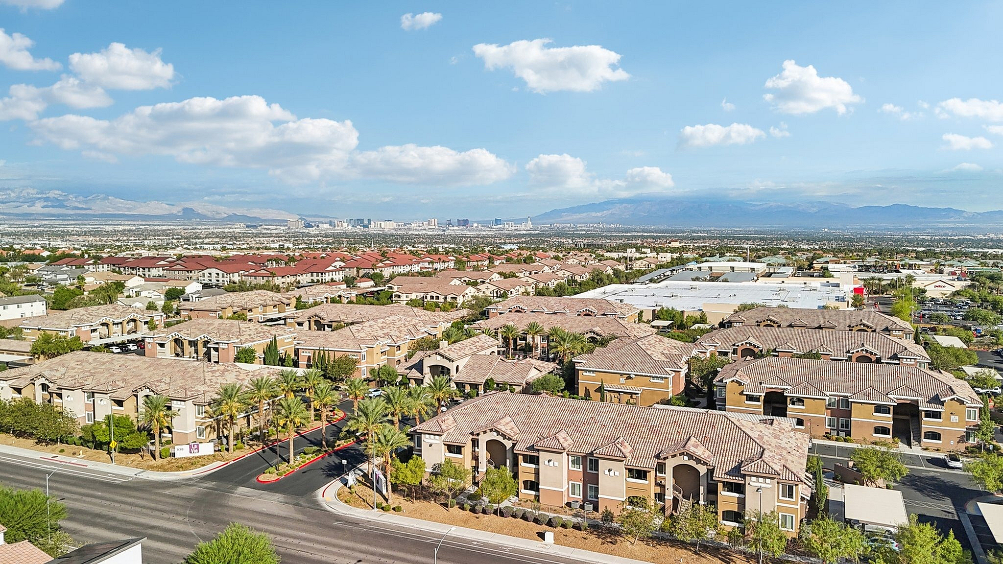 A vibrant aerial view of a modern apartment complex with scenic mountain backdrop, palm-lined streets, and a sprawling cityscape under a clear blue sky.
