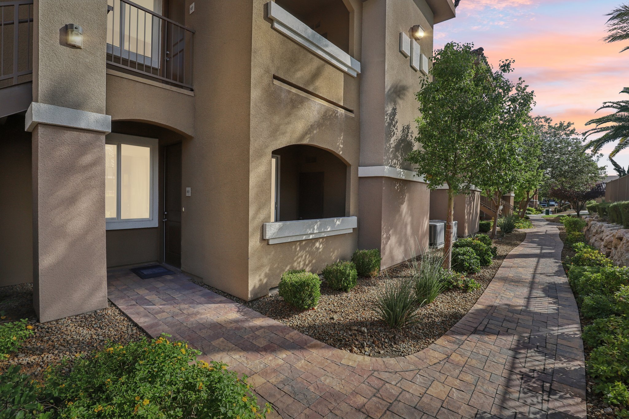 Charming apartment entrance with manicured landscaping and a welcoming footpath under a vibrant sunset sky.
