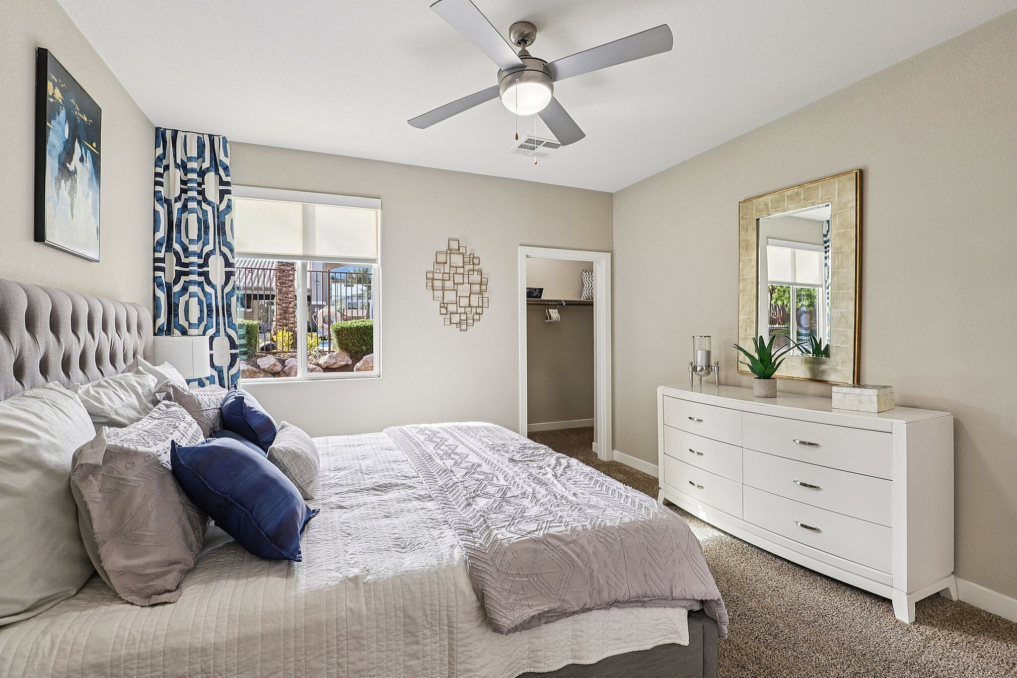 Elegant bedroom with plush bedding, sleek white dresser, and modern decor, inviting natural light through a large window with garden views.