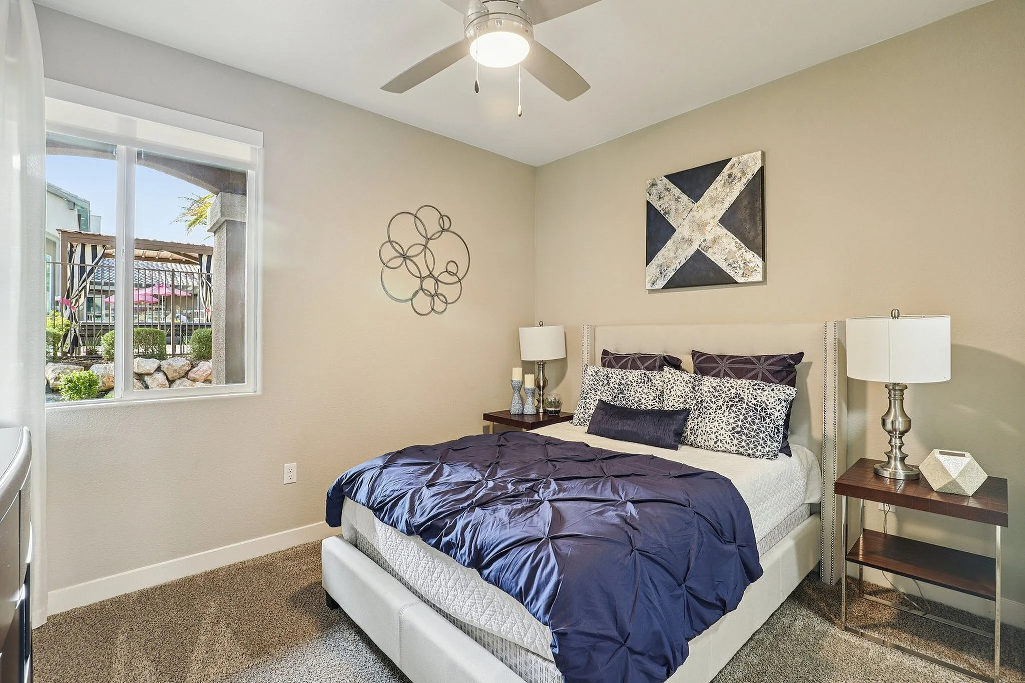 Bright and cozy bedroom featuring a plush bed with elegant blue and white bedding, sleek nightstands, and a stylish ceiling fan, offering a serene view through a large window.