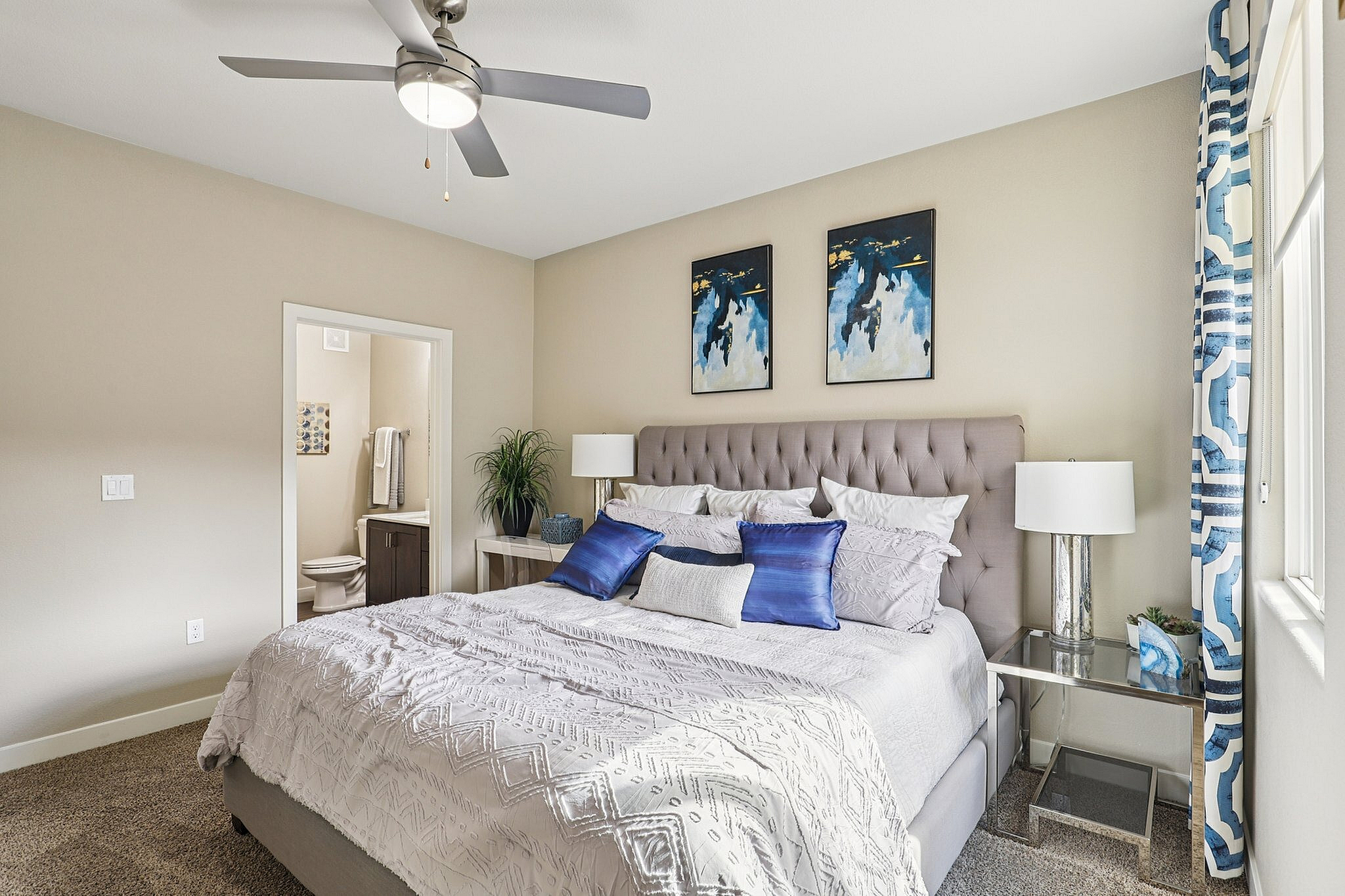 Elegant bedroom featuring a plush bed with blue accent pillows, stylish decor, and natural light from large windows.