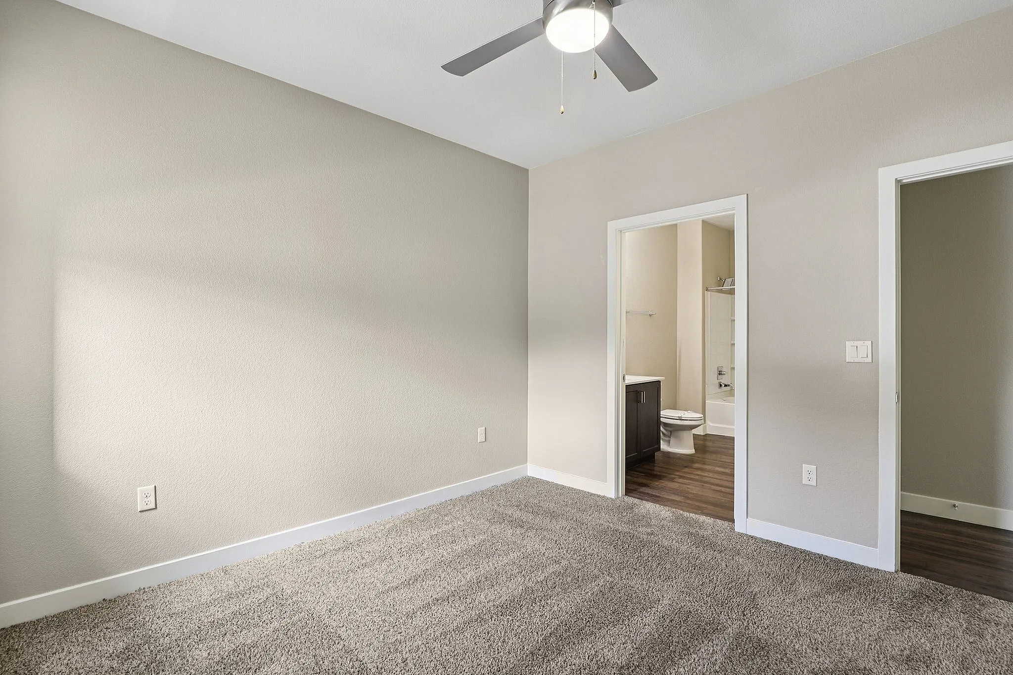 Bright, spacious bedroom with plush carpeting and modern ceiling fan, leading to a sleek en-suite bathroom featuring elegant dark wood cabinetry and contemporary fixtures.