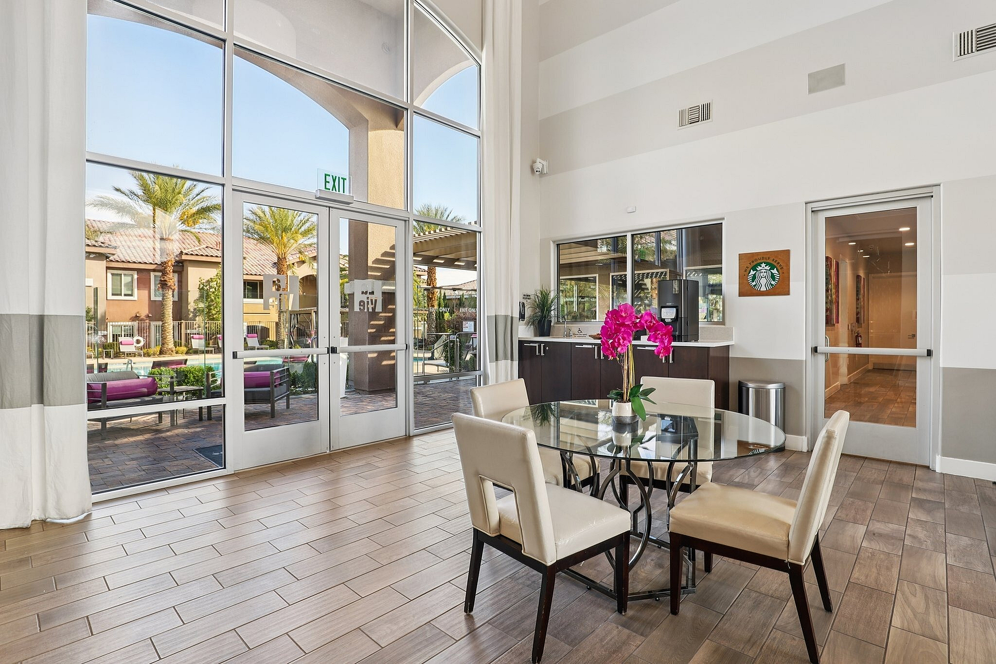 Elegant apartment lobby with high ceilings, large windows, and vibrant decor, offering a bright and inviting atmosphere with views of the palm-lined outdoor patio.