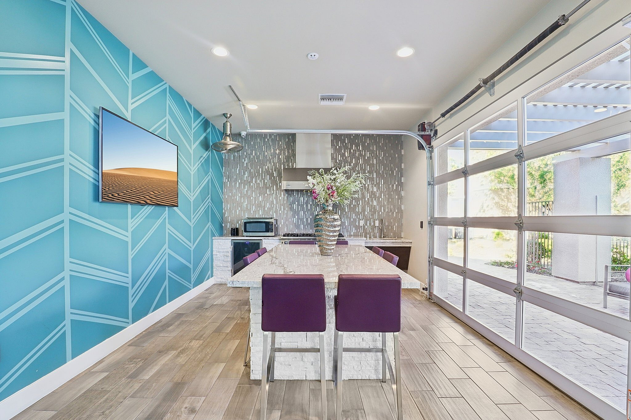 Bright and modern kitchen with sleek tile walls, vibrant blue accent wall, and a spacious island with purple chairs, leading to a sunny outdoor patio.
