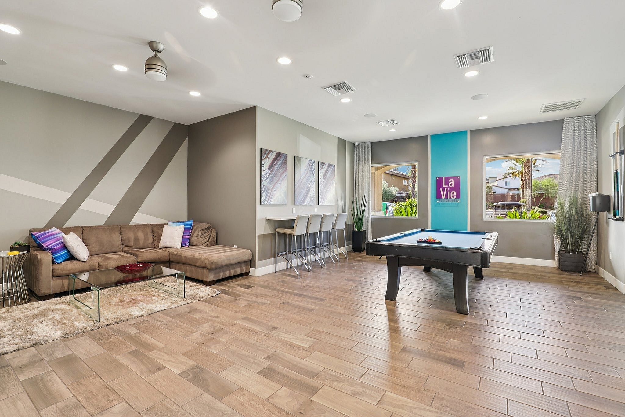 Modern lounge area with cozy sectional sofa and sleek billiards table, enhanced by bright artwork, vibrant cushions, and stylish wood flooring.