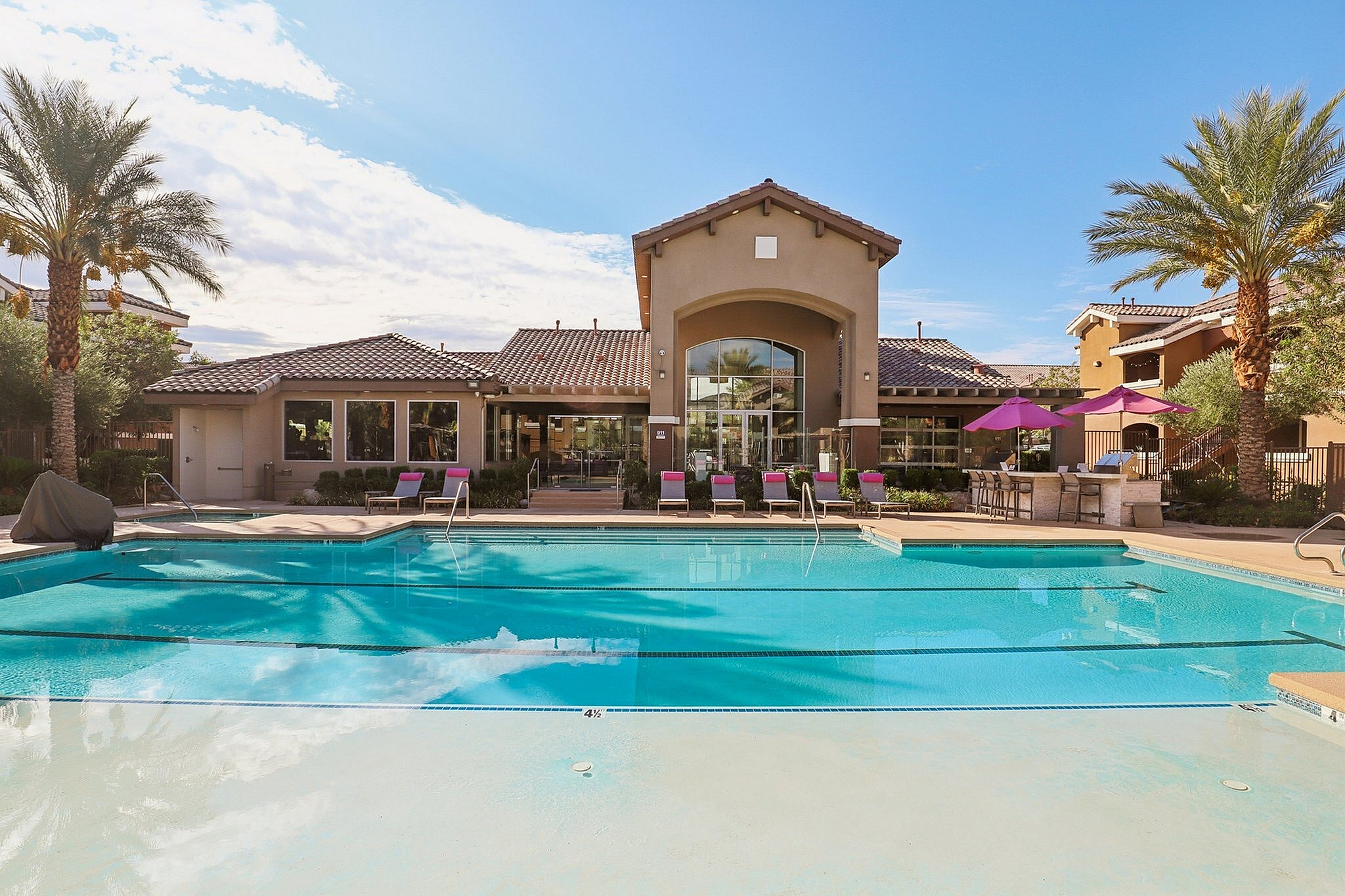 Luxurious apartment complex with a sparkling outdoor pool, modern lounge chairs, and elegant palm trees against a clear blue sky backdrop.