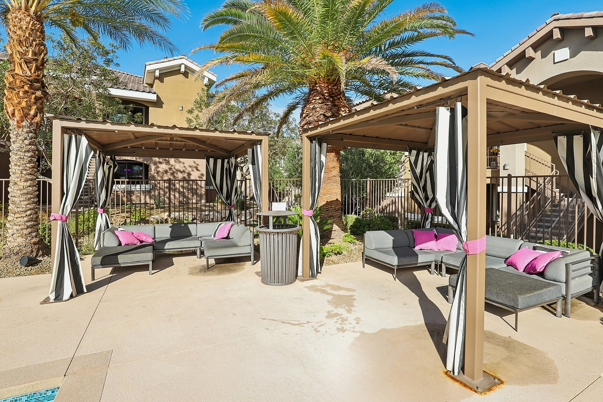 Elegant outdoor lounge area with stylish cabanas featuring chic striped curtains and vibrant pink cushions, surrounded by lush palm trees.