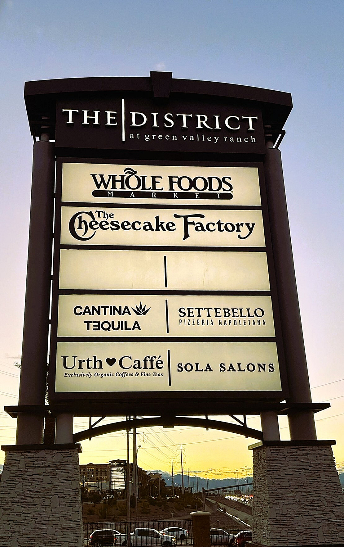 Prominent retail sign for The District at Green Valley Ranch, featuring popular venues like Whole Foods Market, The Cheesecake Factory, and Urth Caffé, set against a scenic sunset skyline.