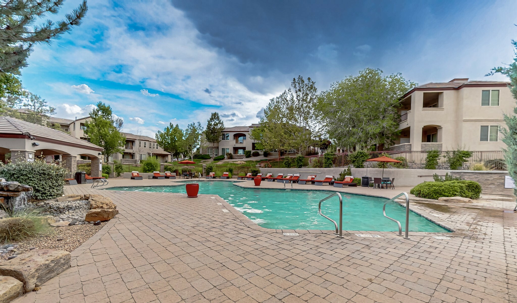 Luxurious apartment complex showcasing a sparkling pool, comfortable lounge chairs, and lush landscaping under a vibrant sky.