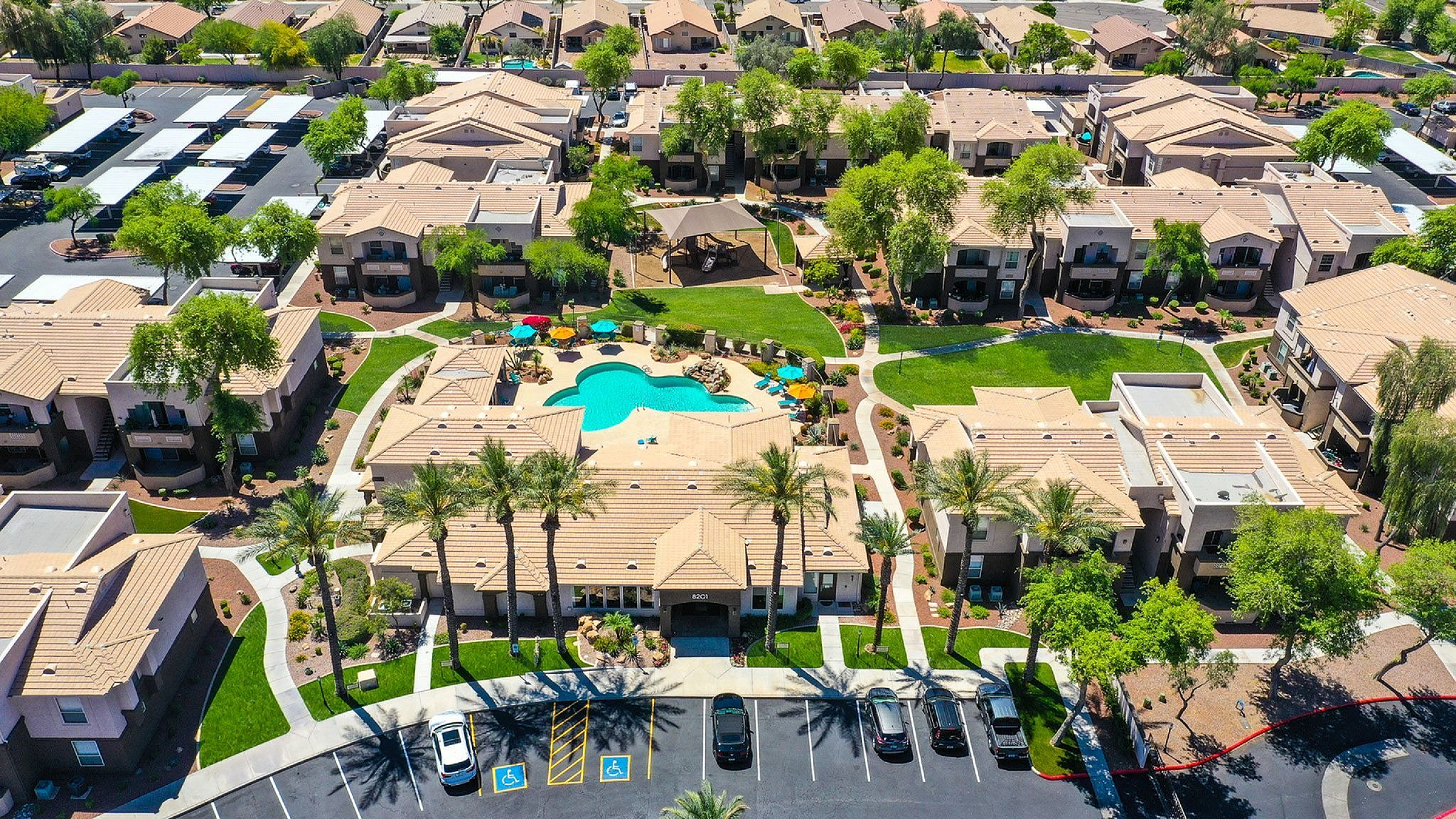Aerial view of a modern apartment complex featuring a vibrant central pool, lush green landscape, and spacious parking area, offering a beautiful and welcoming community environment.