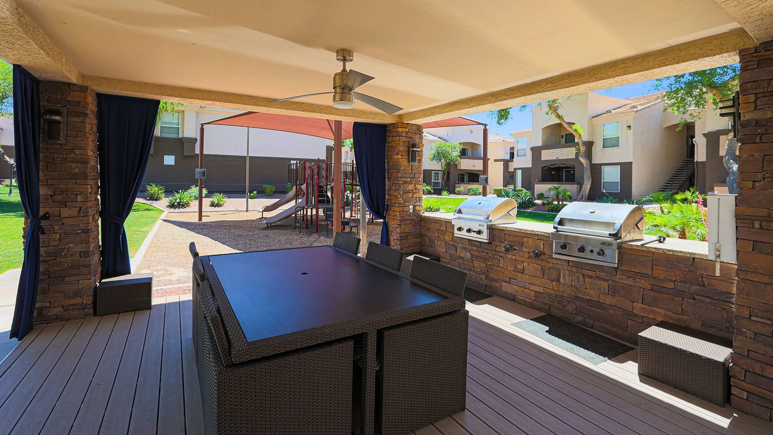 Modern outdoor dining area with built-in grills, ceiling fan, and stylish seating overlooking vibrant apartment community and playground.