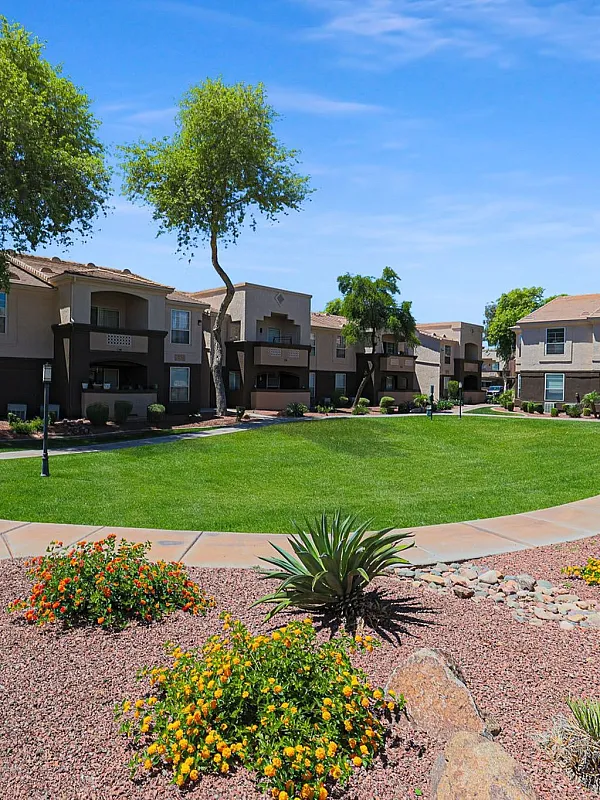 Charming residential community with spacious green lawns, colorful flower beds, and modern two-story apartments under a clear blue sky.