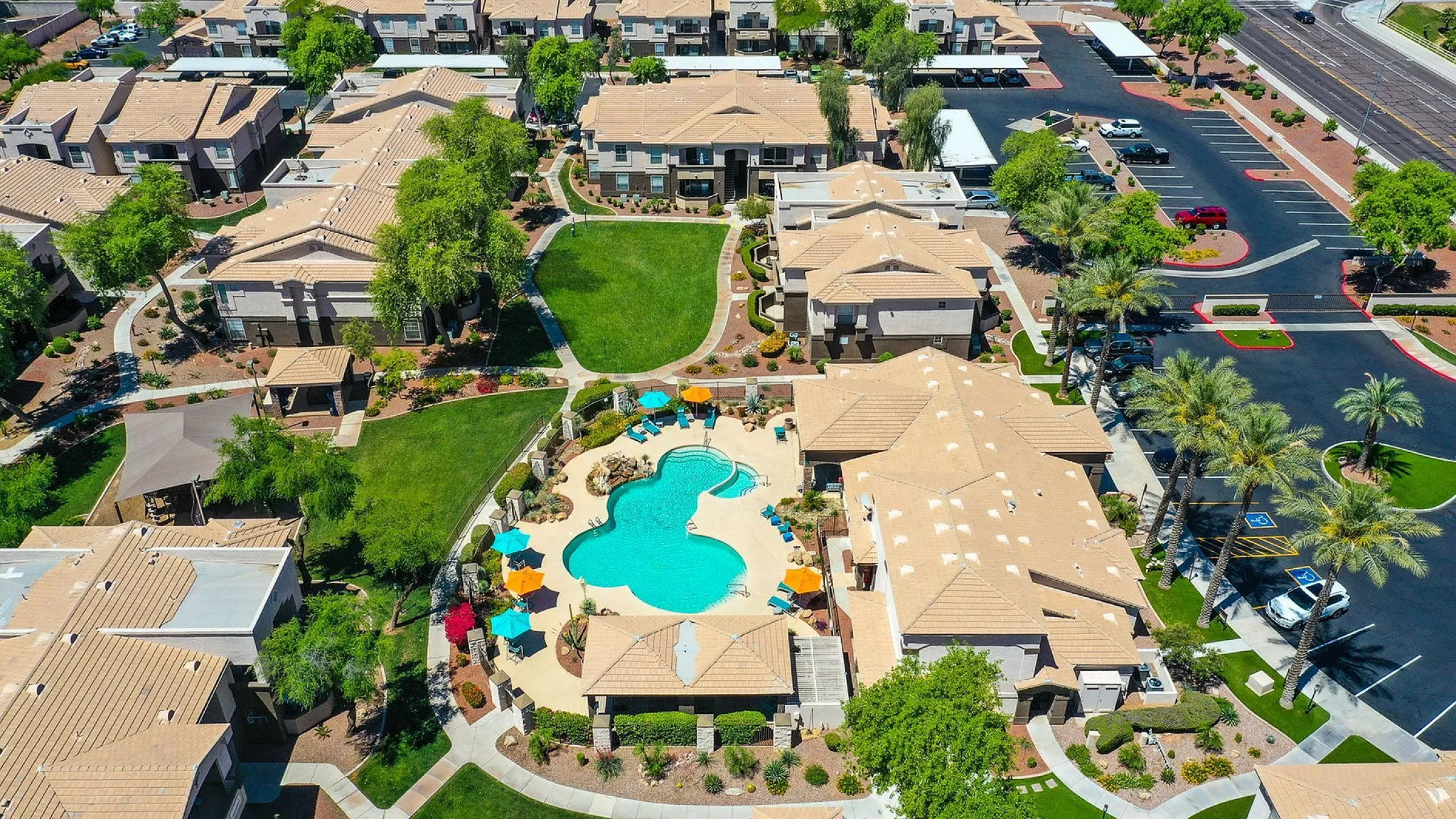 Aerial view of a luxurious apartment complex featuring a resort-style pool with colorful umbrellas, lush green spaces, and well-manicured landscaping.
