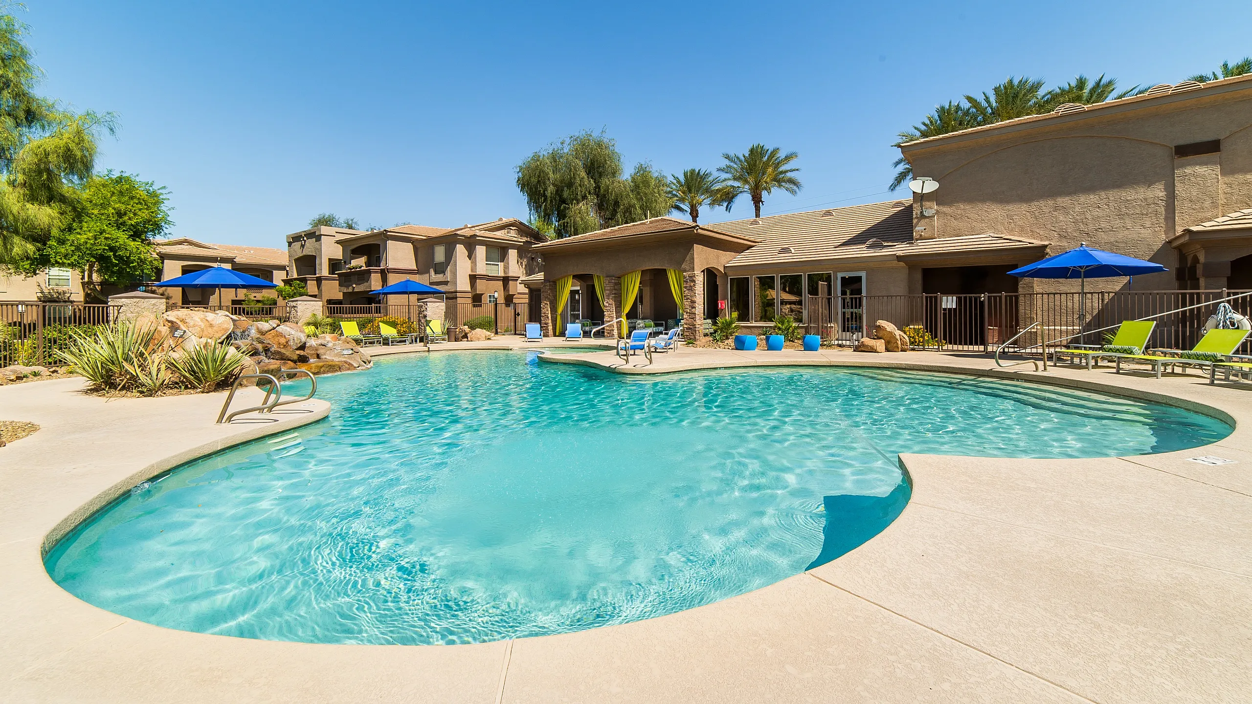 Luxurious apartment complex showcasing a sparkling resort-style swimming pool, surrounded by lush landscaping and modern amenities under a clear blue sky.