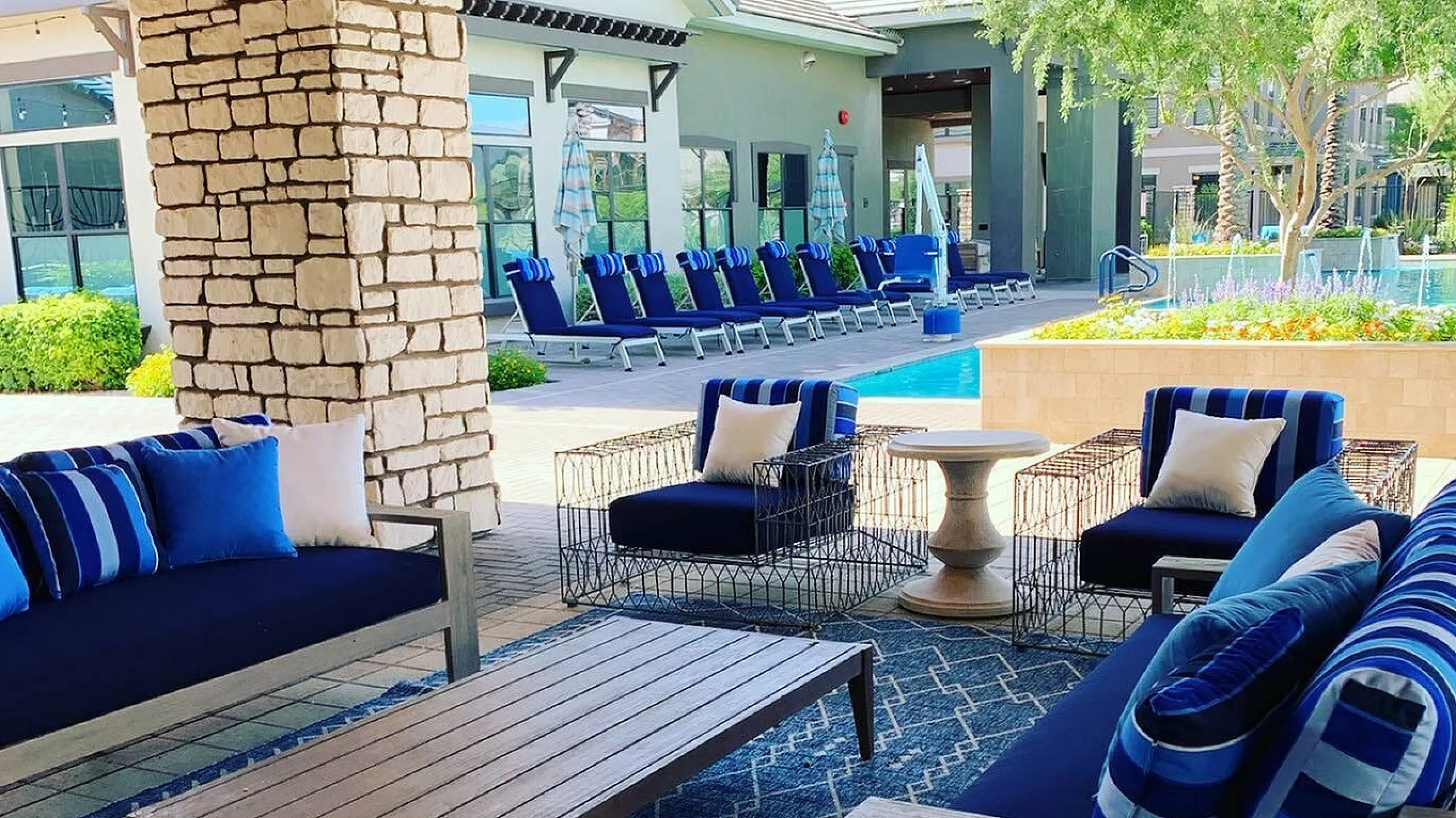 Chic outdoor seating area with stylish navy blue cushions, overlooking a pristine pool and sun loungers, set in a lush green apartment complex courtyard.